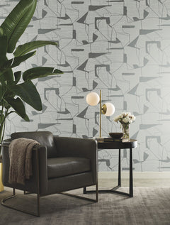 A modern living space featuring a gray armchair, a round black side table with a lamp and flowers, a large leafy plant, and York Wallcoverings’ Abstract Geo Wallpaper in the background.