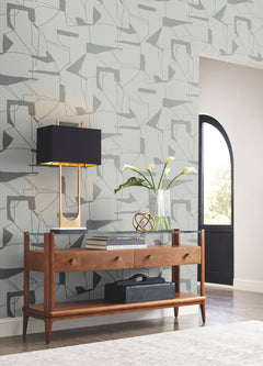 A wooden console table with two drawers stands against a wall adorned with York Wallcoverings’ Abstract Geo Wallpaper. On the table are a dark lamp, a glass vase holding white flowers, and a glass sculpture. Sunlight enters through an arched doorway.