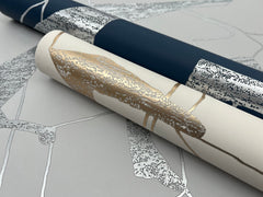 Two rolls of York Wallcoverings’ Abstract Geo Wallpaper—one in dark blue with a silver motif and the other in cream featuring gold and silver patterns—rest on a flat surface adorned with wallpaper showcasing a silver abstract leaf design.