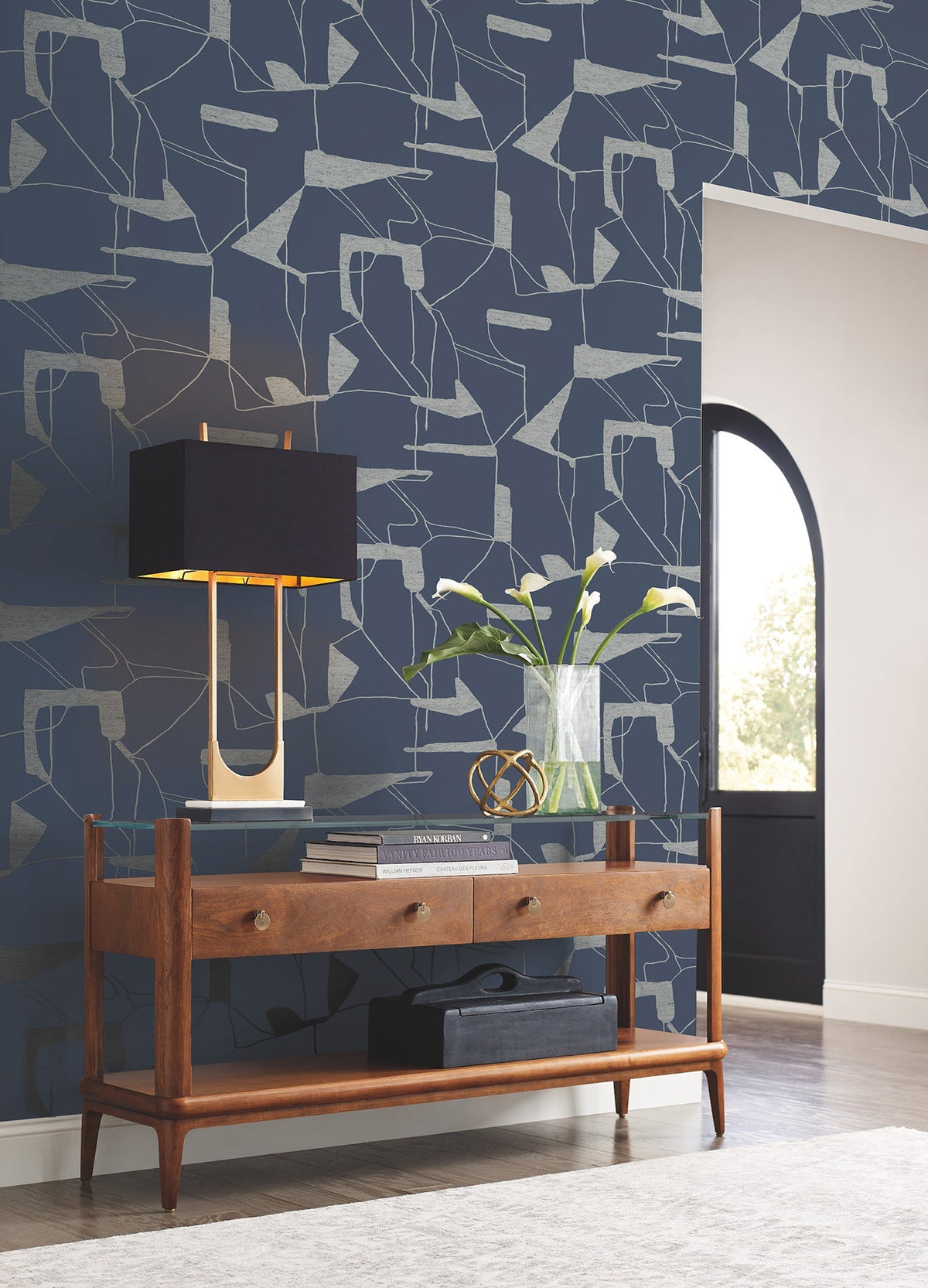A mid-century modern console table with drawers holds a black and gold lamp, a vase with white flowers, and decorative items. The wall behind features Abstract Geo Wallpaper by York Wallcoverings in navy, while an arched doorway reveals sunlight outside.