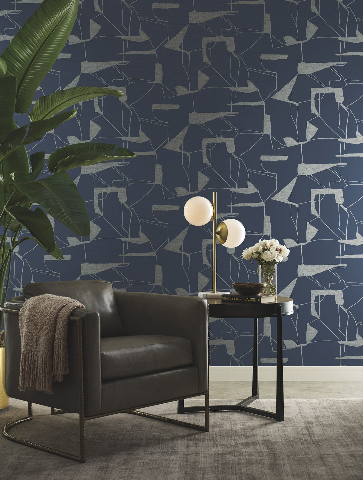 A modern living space featuring a dark leather armchair, a side table with a lamp and flowers, a large plant, and York Wallcoverings’ Abstract Geo Wallpaper in blue. A neutral rug covers the floor.