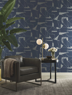 A modern living space featuring a dark leather armchair, a side table with a lamp and flowers, a large plant, and York Wallcoverings’ Abstract Geo Wallpaper in blue. A neutral rug covers the floor.