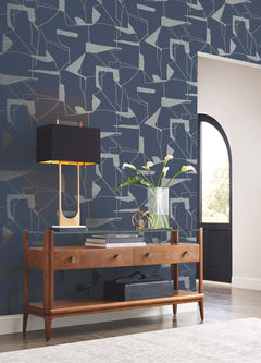 A mid-century console table with a glass top holds a black lamp, a glass vase with white tulips, and a decorative sphere. Behind it, the wall is adorned with Abstract Geo Wallpaper by York Wallcoverings featuring a navy geometric pattern.