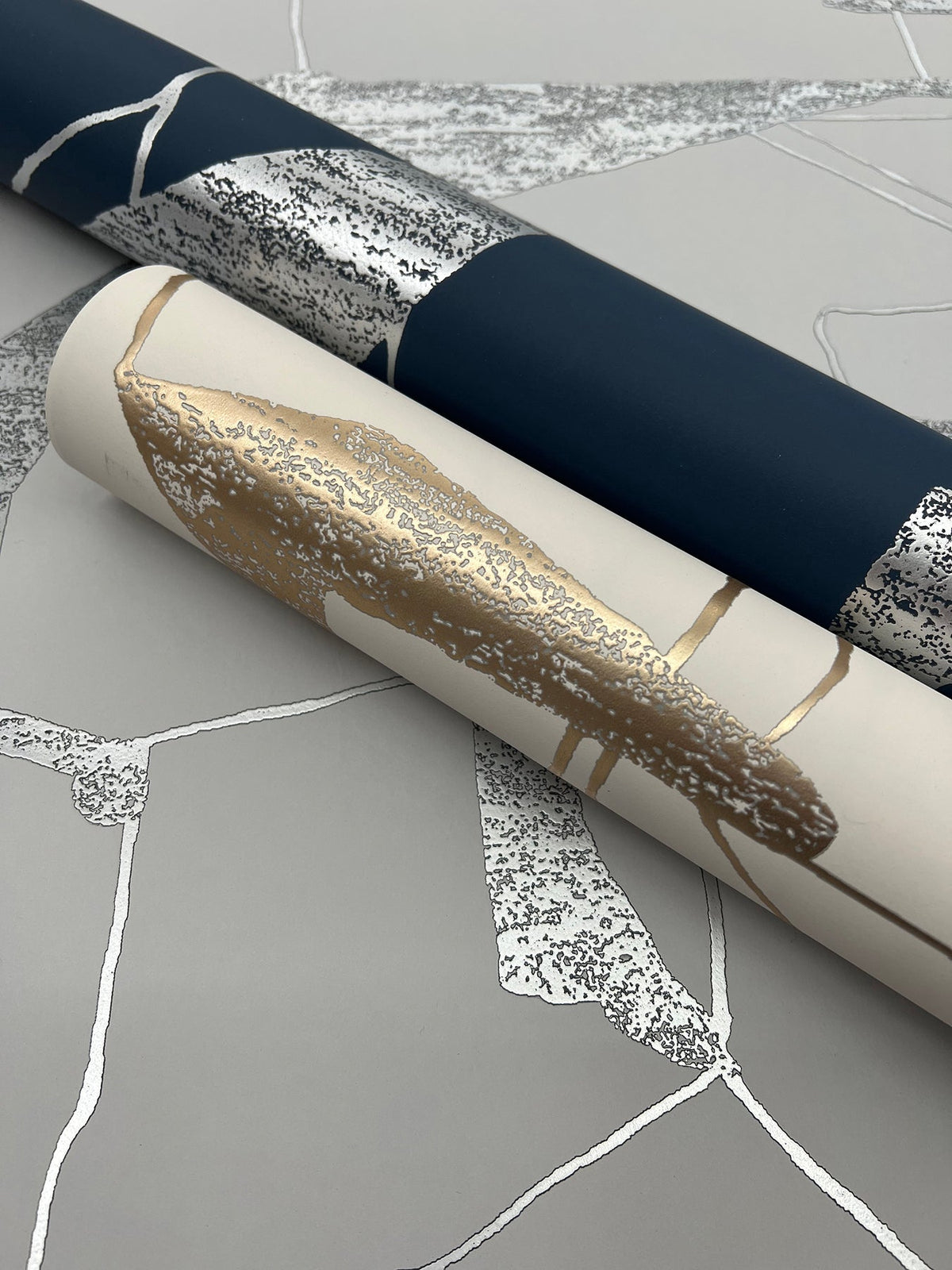 Two rolls of York Wallcoverings’ Abstract Geo Wallpaper—one in a dark blue and silver abstract pattern, and the other featuring a white, gold, and silver design—are arranged on a flat gray surface displaying a coordinated abstract motif.