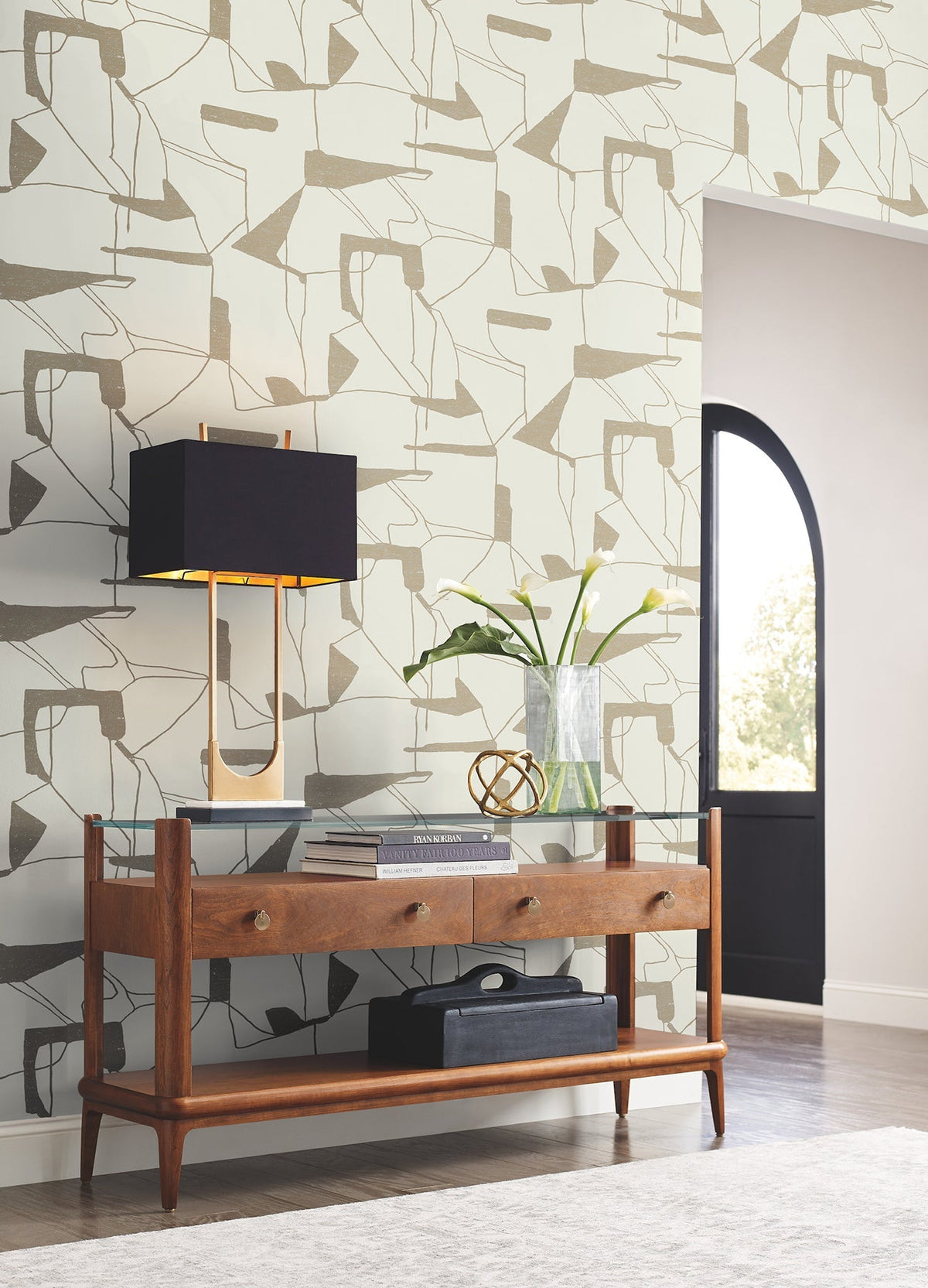 Modern entryway featuring York Wallcoverings’ Abstract Geo Wallpaper, a wooden console table with two drawers, a black and gold wall lamp, a glass vase filled with green branches, and a round glass decor piece. Light streams in from an arched doorway.