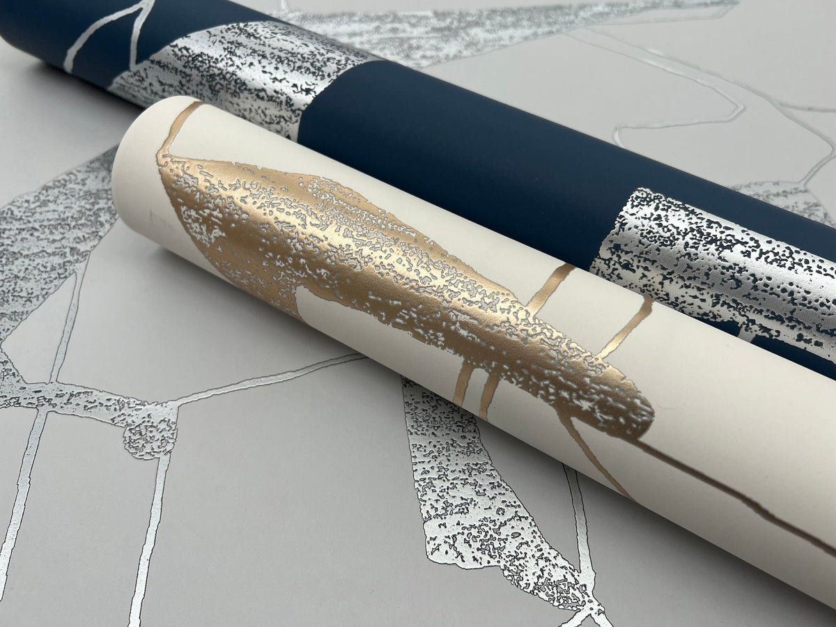 Two rolls of York Wallcoverings Abstract Geo Wallpaper—one in dark blue with silver accents, the other in white with gold—rest on a flat surface covered in a silver and gray leaf-patterned wallpaper. The textures and metallic details of the Abstract Geo designs are clearly visible.