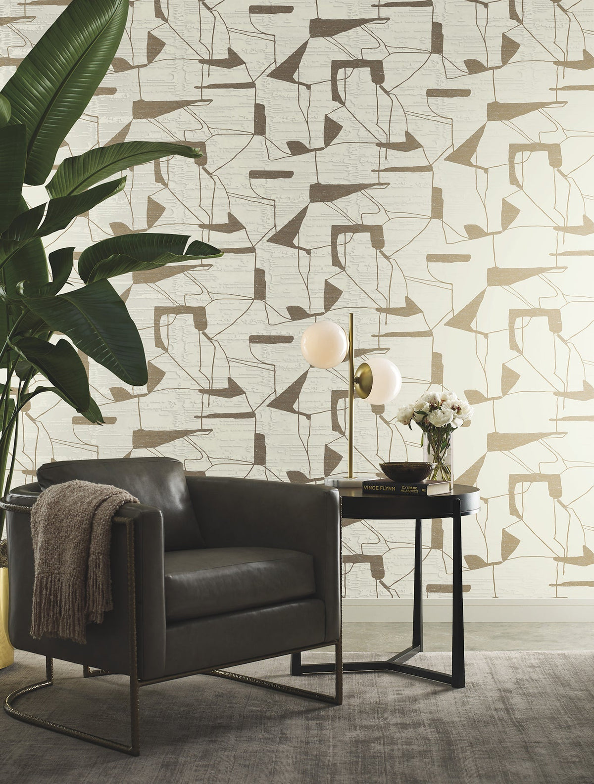 A modern reading nook featuring a gray armchair, a round black side table, a globe lamp, a small bouquet of flowers, a throw blanket, a large plant, and York Wallcoverings’ Abstract Geo Wallpaper in beige.
