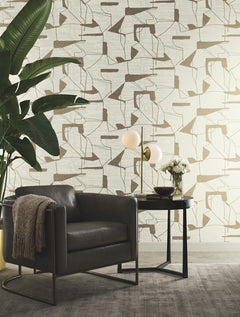A modern reading nook featuring a gray armchair, a round black side table, a globe lamp, a small bouquet of flowers, a throw blanket, a large plant, and York Wallcoverings’ Abstract Geo Wallpaper in beige.