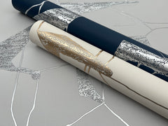 Two rolls of York Wallcoverings Abstract Geo Wallpaper—one in navy with silver accents and the other in cream with gold accents—are placed on a flat surface, showcasing their striking geometric pattern in silver and gray.