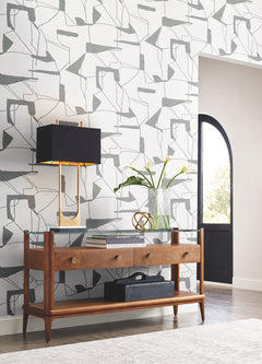A modern entryway features a wooden console table topped with a black lamp, glass vase of white flowers, and decorative accents. Sunlight pours in through an arched doorway, highlighting the Abstract Geo Wallpaper by York Wallcoverings in the background.