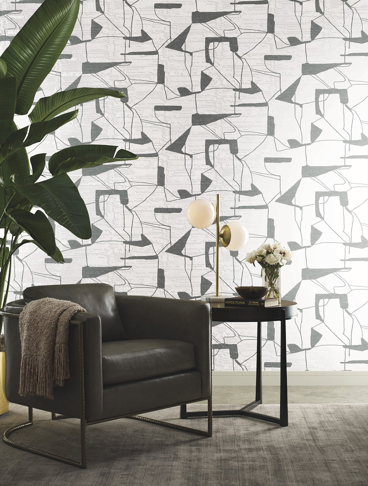 Modern living space with a gray leather armchair, a small round table holding a lamp and flowers, a large green potted plant, and Abstract Geo Wallpaper by York Wallcoverings in the background.