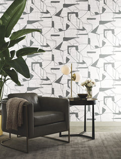 Modern living space with a gray leather armchair, a small round table holding a lamp and flowers, a large green potted plant, and Abstract Geo Wallpaper by York Wallcoverings in the background.