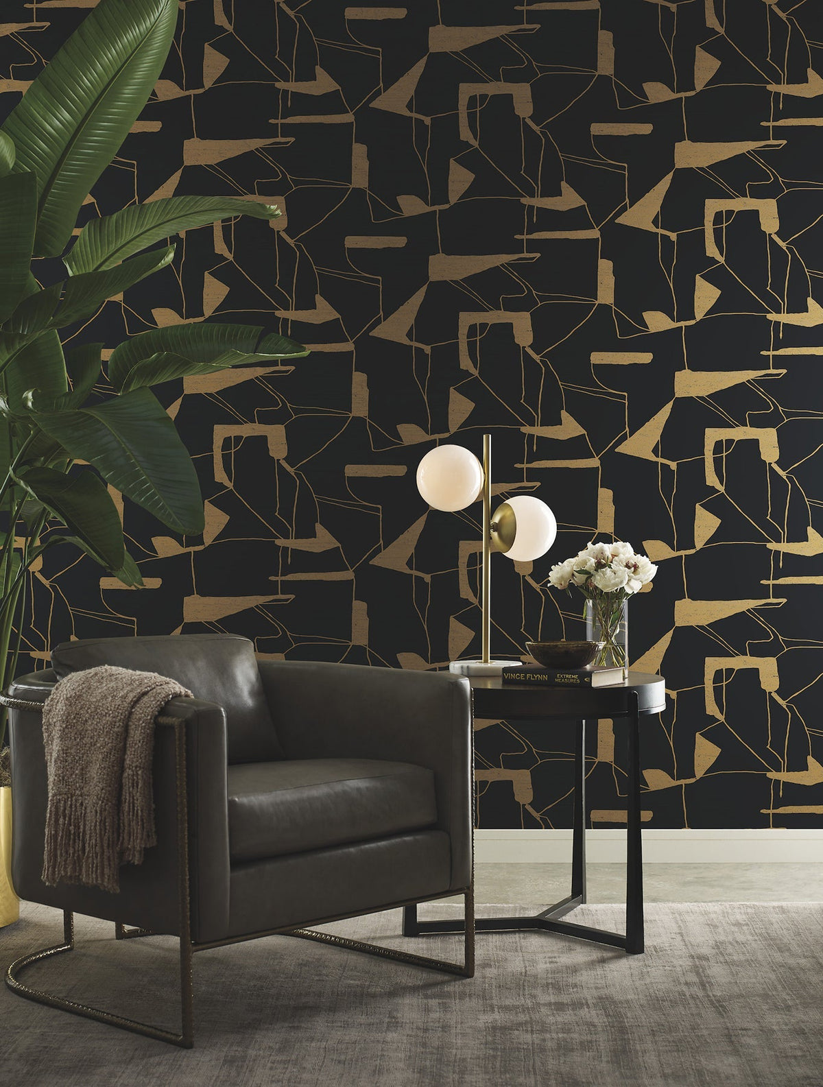 Modern living room featuring York Wallcoverings’ Abstract Geo Wallpaper in black and gold, a dark leather armchair with a draped blanket, a side table displaying flowers and a globe lamp, and a tall green plant in the corner.