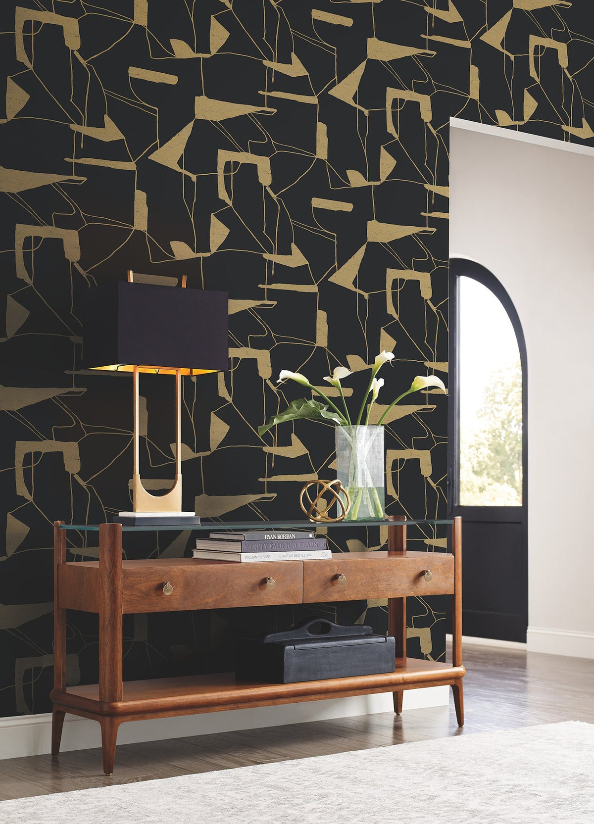 Mid-century console table with two drawers, black lamp, vase with white flowers, and decorative objects are arranged against York Wallcoverings’ Abstract Geo Wallpaper in black and gold. Sunlight streams through a nearby arched doorway onto the floor.