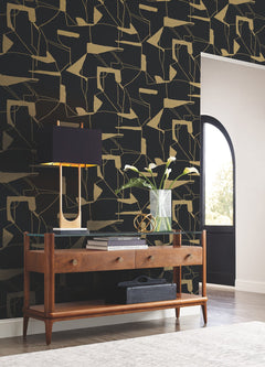 Mid-century console table with two drawers, black lamp, vase with white flowers, and decorative objects are arranged against York Wallcoverings’ Abstract Geo Wallpaper in black and gold. Sunlight streams through a nearby arched doorway onto the floor.