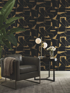 A modern living space featuring York Wallcoverings’ Abstract Geo Wallpaper in a dark geometric design, complemented by a black leather chair, a tall green plant, a side table with flowers, and a contemporary lamp—all set atop a light gray rug.