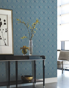 A modern room featuring Harlowe Wallpaper by York Wallcoverings in a blue pattern, complemented by a black console table, a vase with yellow flowers, a bowl of fruit, wall art, and a beige chair positioned by a window with sheer curtains.