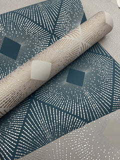 Rolls of Harlowe Wallpaper by York Wallcoverings featuring a geometric starburst pattern in metallic silver and gold on teal and beige backgrounds, arranged overlapping each other.