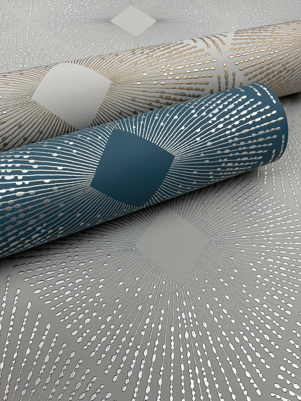 Two rolls of Harlowe Wallpaper by York Wallcoverings—one in teal and one in beige—featuring metallic dotted lines radiating from diamond shapes, are displayed on a matching patterned surface.