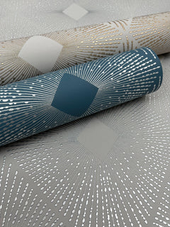 Two rolls of Harlowe Wallpaper by York Wallcoverings—one in teal and one in beige—featuring metallic dotted lines radiating from diamond shapes, are displayed on a matching patterned surface.