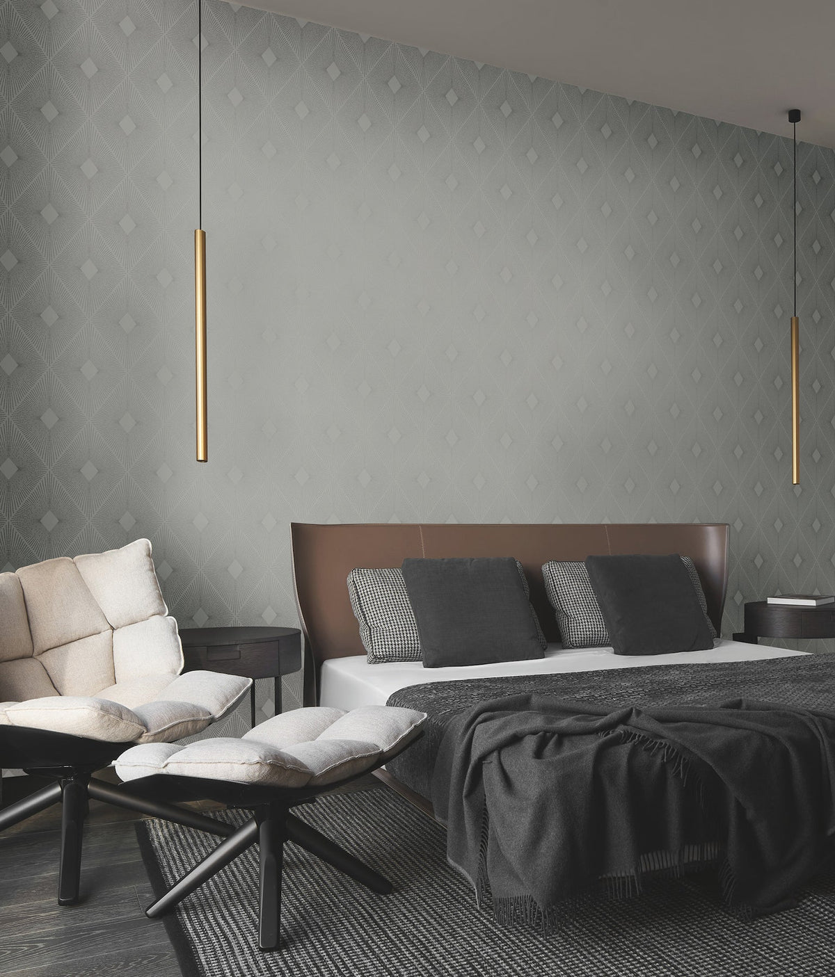 Modern bedroom featuring Harlowe Wallpaper by York Wallcoverings in a gray geometric pattern, a brown leather headboard, gray bedding, a cream sofa, two round ottomans, two hanging cylindrical gold lights, and dark wood flooring.