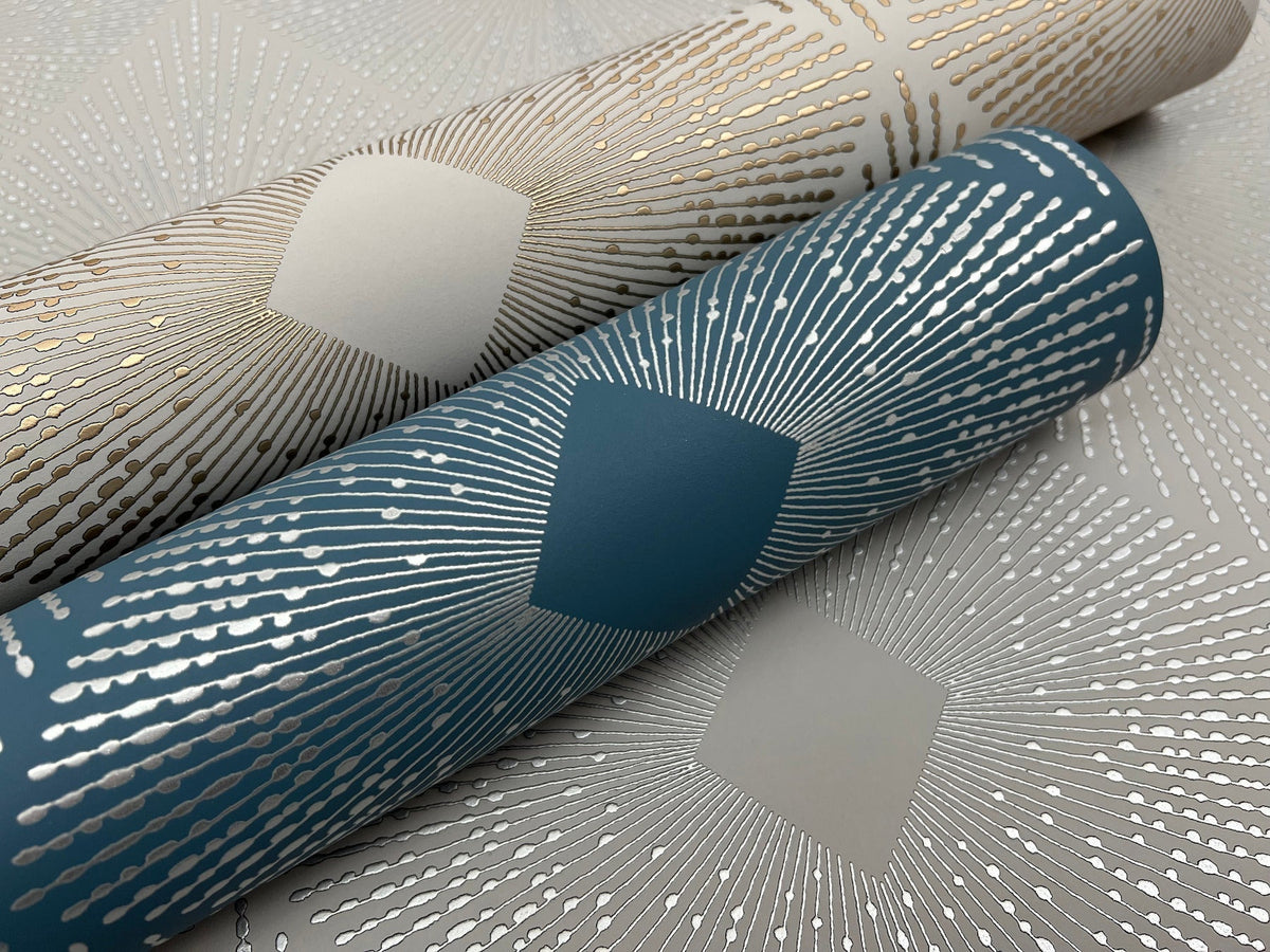 Two rolls of Harlowe Wallpaper by York Wallcoverings—one in teal with silver geometric sunburst patterns and the other in beige with gold sunburst designs—are displayed on a matching patterned wallpaper backdrop.
