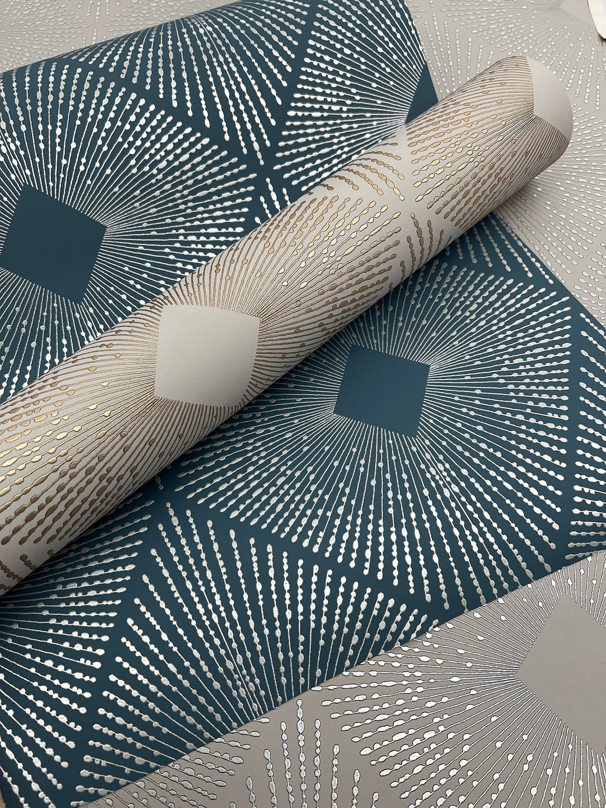A close-up of York Wallcoverings’ Harlowe Wallpaper showcases its blue and beige geometric patterns with metallic silver accents, featuring radiating lines and diamond shapes. A roll of the Harlowe Wallpaper is partially unrolled on top.