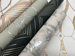 Four rolls of Shimmering Foliage Wallpaper by York Wallcoverings in beige, black, and gray hues—each featuring metallic gold or silver geometric and floral designs—are arranged diagonally on a white and gold-patterned surface.