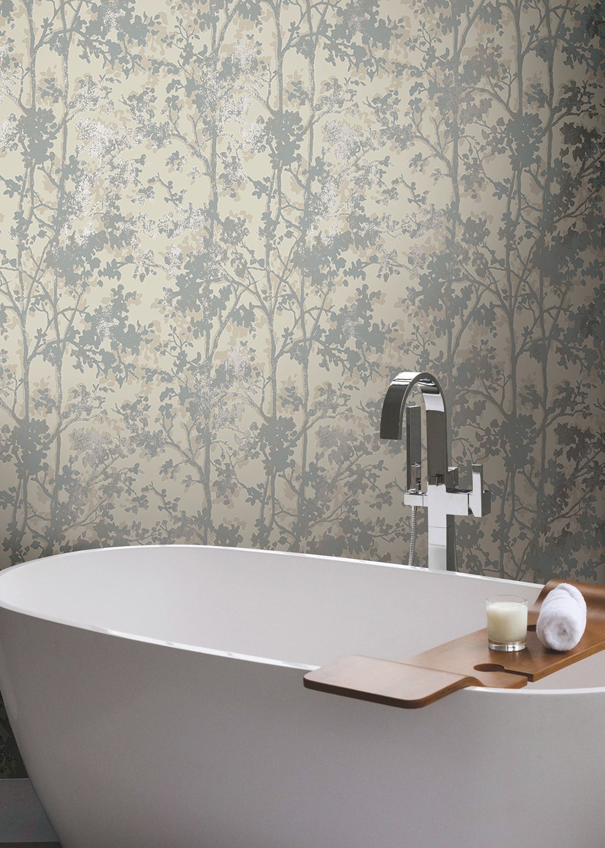 A modern white bathtub with a wooden tray holding a candle and towel sits in front of York Wallcoverings’ Shimmering Foliage Wallpaper, featuring a subtle tree and leaf pattern in soft beige and gray tones.