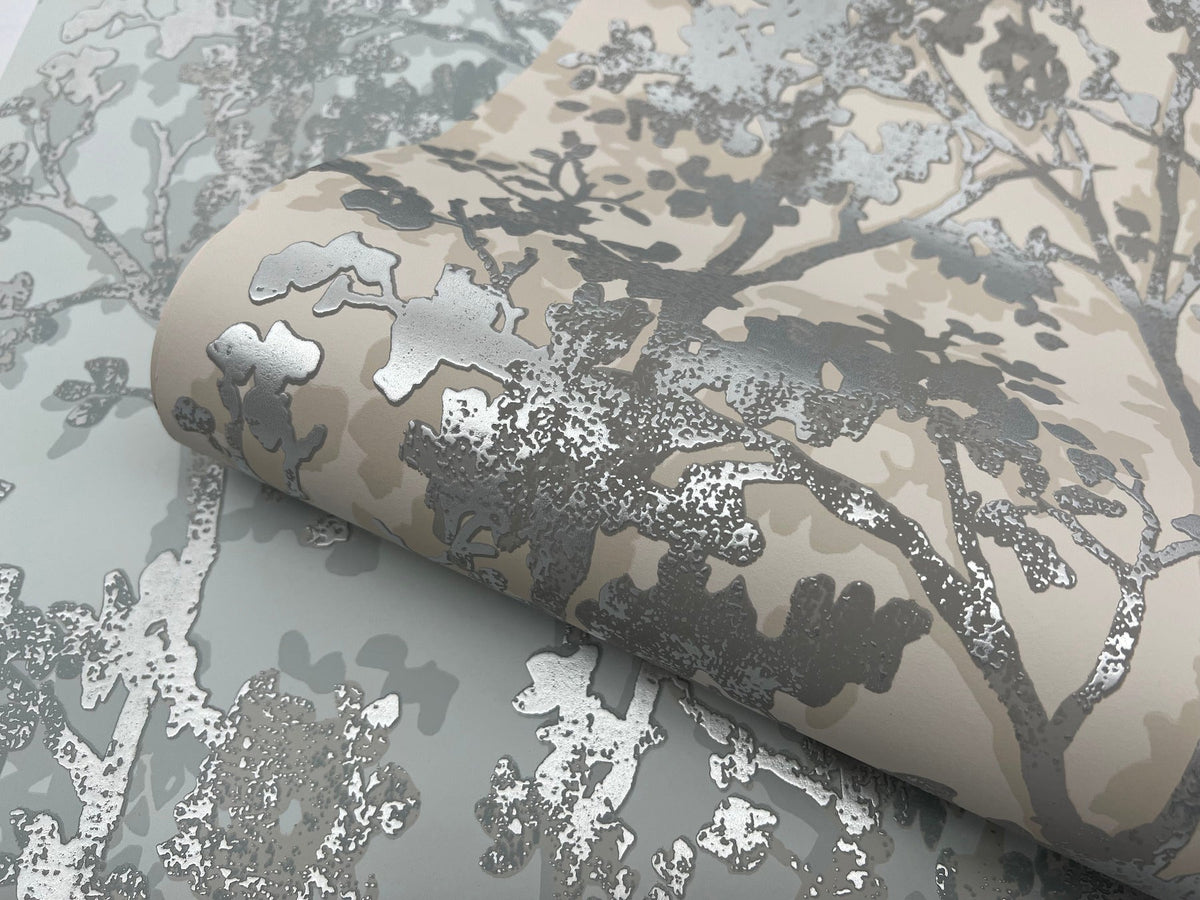 A partially unrolled roll of York Wallcoverings’ Shimmering Foliage Wallpaper, featuring a metallic silver tree branch motif on a beige and light gray background, displayed on a matching surface.