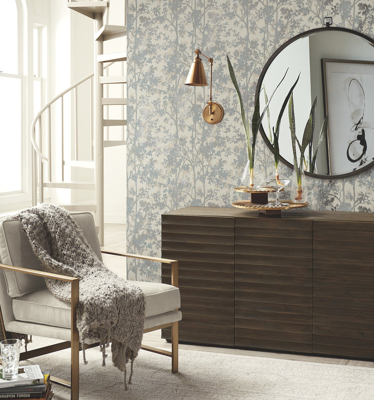 Modern living room featuring a velvet armchair, knitted throw, spiral staircase, wooden sideboard, round mirror, tall plants, and soft-toned Shimmering Foliage Wallpaper by York Wallcoverings. Art and books add decorative touches.