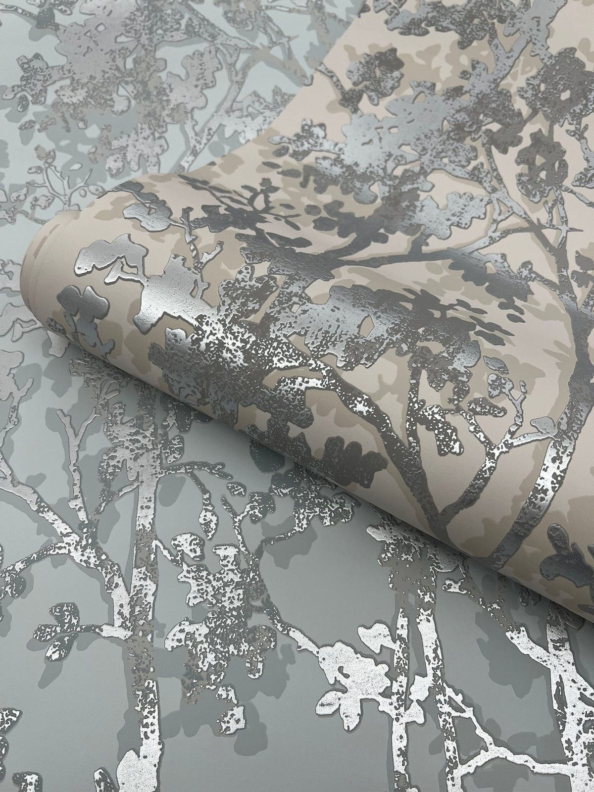 A close-up of York Wallcoverings’ Shimmering Foliage Wallpaper, featuring metallic silver and beige branches and leaves on a light grey background, with one section rolled up atop the flat surface.