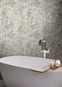A modern white bathtub with a wooden tray holding a candle and rolled towel sits in front of York Wallcoverings’ Shimmering Foliage Wallpaper, which features a subtle leafy tree pattern in soft, neutral tones.