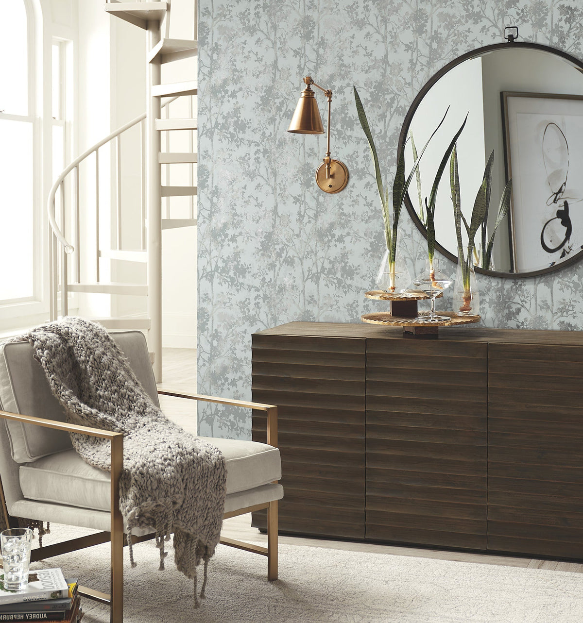 A modern living room featuring York Wallcoverings’ Shimmering Foliage Wallpaper, a beige armchair draped with a gray knit throw, wood sideboard, tall plants, round mirror, abstract art, and a spiral staircase in the background.