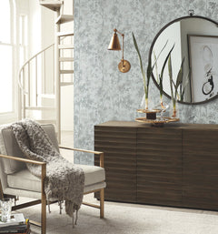 A modern living room featuring York Wallcoverings’ Shimmering Foliage Wallpaper, a beige armchair draped with a gray knit throw, wood sideboard, tall plants, round mirror, abstract art, and a spiral staircase in the background.