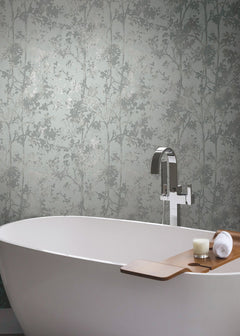 A modern white bathtub with a wooden tray holding a candle and towel is set against York Wallcoverings’ Shimmering Foliage Wallpaper, complemented by a sleek silver faucet.