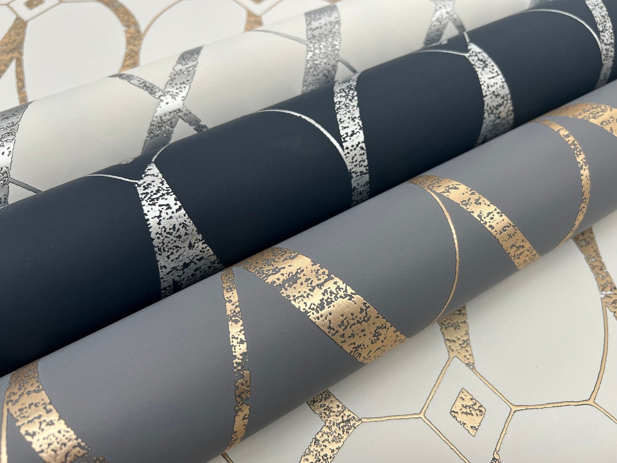 Three rolls of Graceful Geo Wallpaper by York Wallcoverings, featuring metallic geometric patterns in silver and gold on dark blue, gray, and off-white backgrounds, are displayed on a coordinating patterned surface.