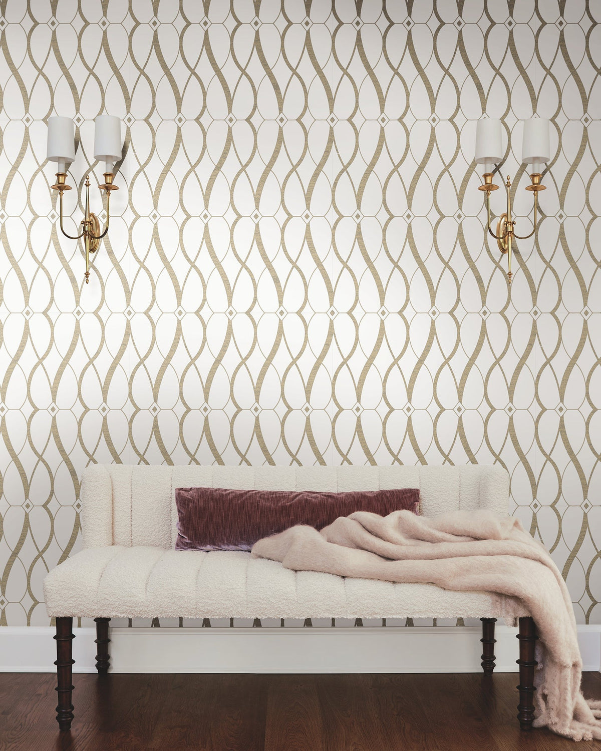 A wall adorned with York Wallcoverings’ Graceful Geo Wallpaper in a cream and gold pattern features two wall sconces above a cream upholstered bench. A mauve pillow and a soft beige throw blanket rest on the bench, which sits atop a dark wood floor.