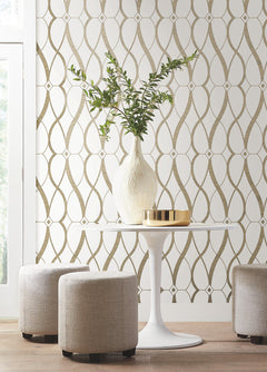 A round white table with a tall vase of greenery and a gold tray, surrounded by three beige ottomans, set against York Wallcoverings’ Graceful Geo Wallpaper in modern beige and white tones.