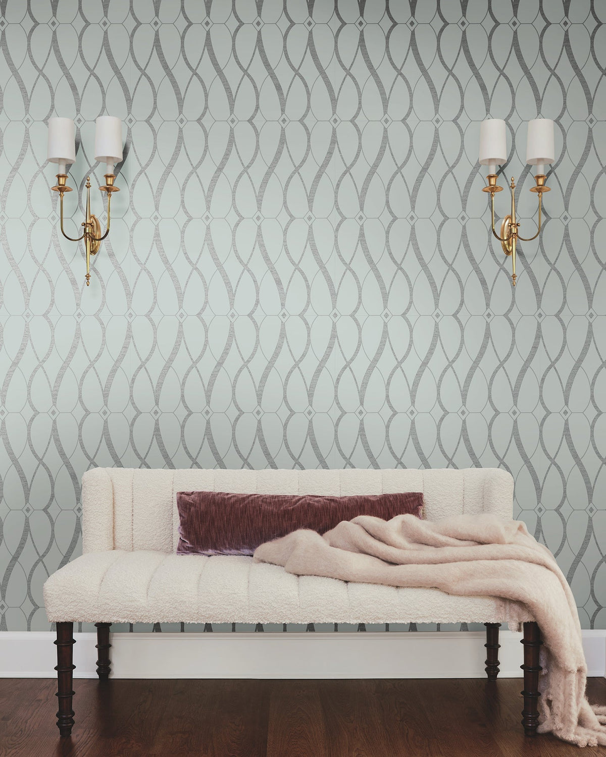 A cream-colored bench with dark wooden legs is topped with a mauve pillow and a soft beige throw. In the background, the walls are adorned with *Graceful Geo Wallpaper* by York Wallcoverings and flanked by two gold wall sconces with white shades.