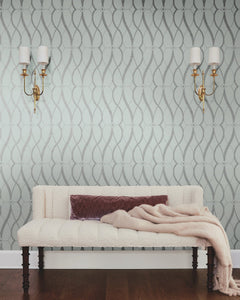 A cream bench with a mauve pillow and blanket sits against a wall adorned with Graceful Geo Wallpaper by York Wallcoverings. Two gold wall sconces with white shades hang on either side above the bench, while the floor is dark wood.