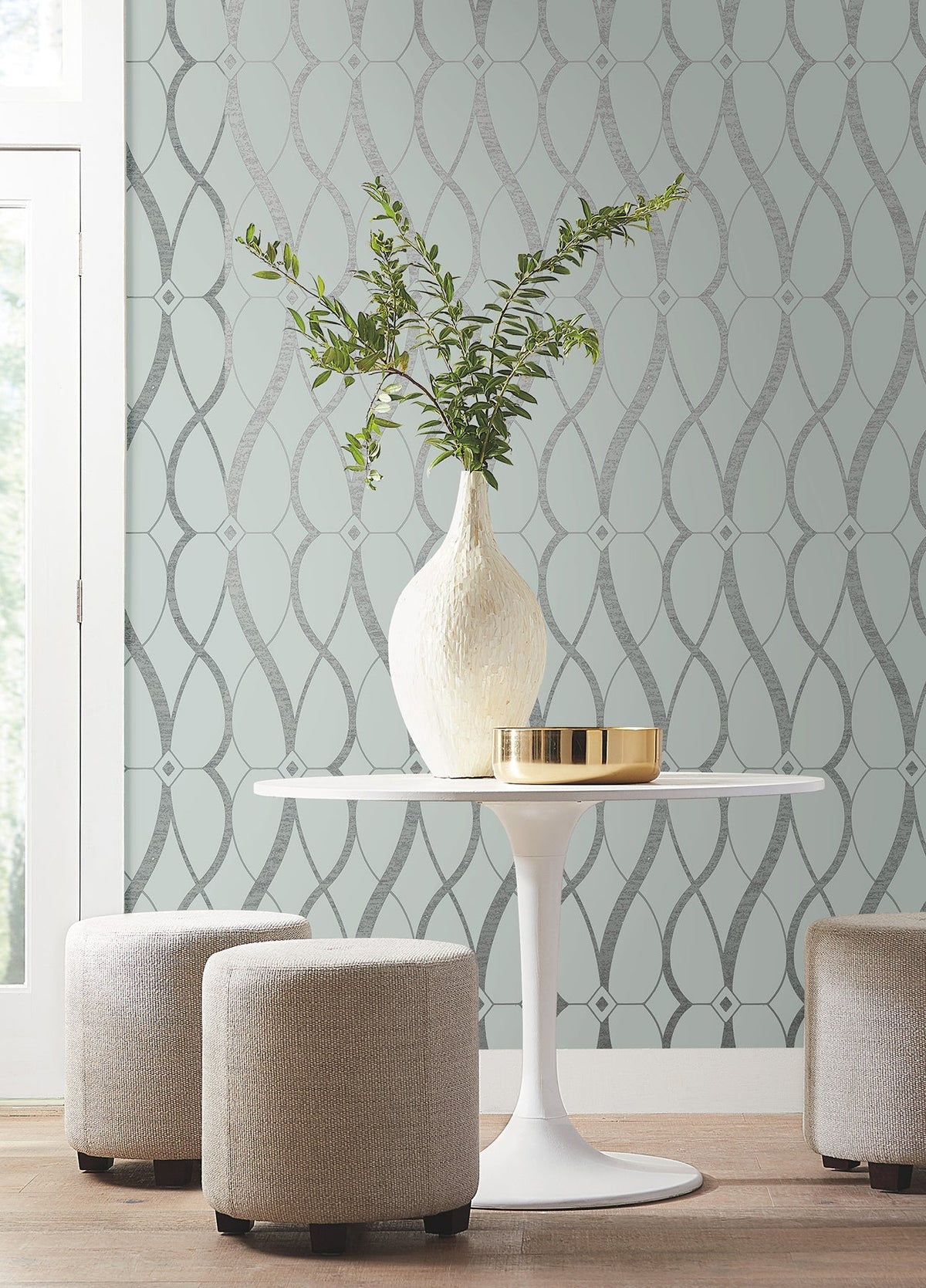 A round white table with a white vase and greenery sits between three beige ottomans. The background features York Wallcoverings’ Graceful Geo Wallpaper in light blue with a silver geometric pattern. Natural light streams in from a window on the left.