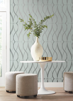 A round white table with a white vase and greenery sits between three beige ottomans. The background features York Wallcoverings’ Graceful Geo Wallpaper in light blue with a silver geometric pattern. Natural light streams in from a window on the left.