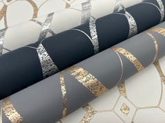 Three rolls of Graceful Geo Wallpaper by York Wallcoverings, each featuring a geometric pattern with metallic silver and gold accents on white, dark navy, and gray backgrounds. One roll is partially unrolled underneath the others.
