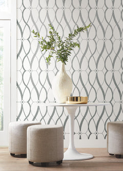 A round white table with a white vase and gold tray sits between three light gray ottomans. The background features York Wallcoverings’ Graceful Geo Wallpaper, showcasing a modern geometric pattern in gray and white. Natural light streams in from a window on the left.