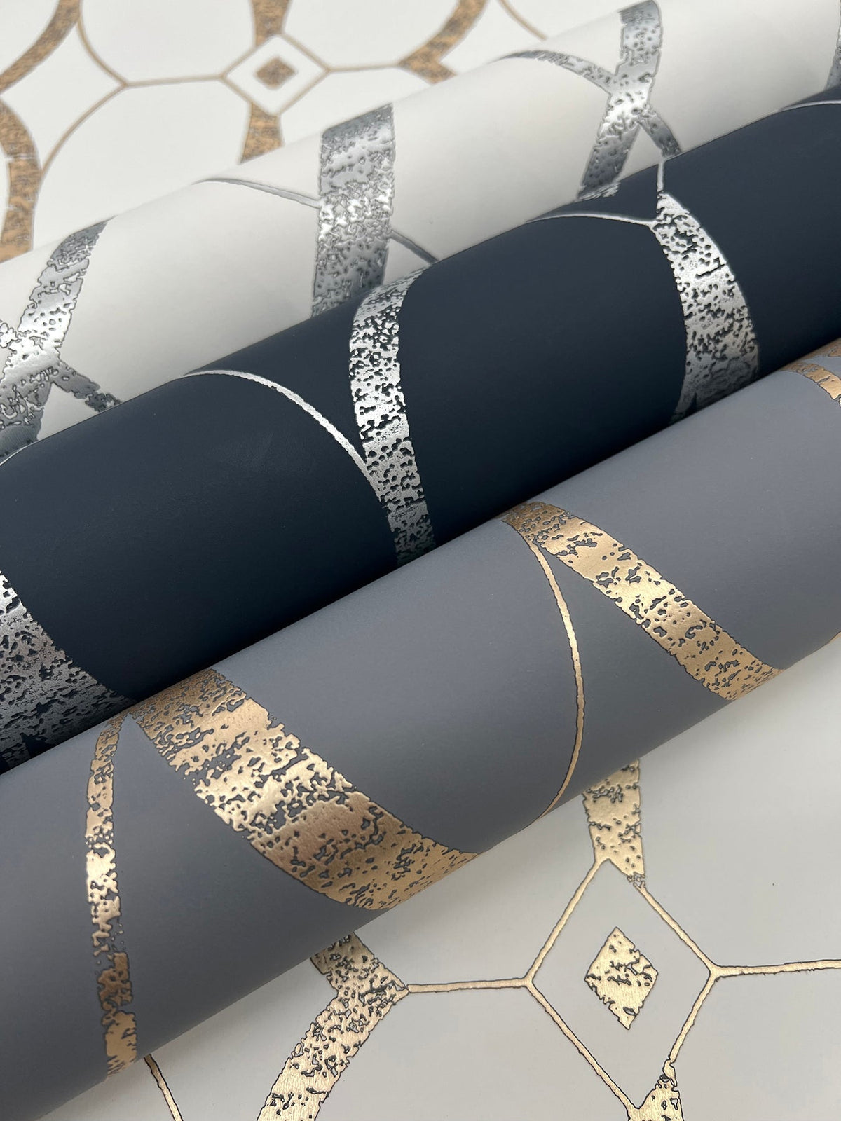 Three rolls of Graceful Geo Wallpaper by York Wallcoverings, each featuring metallic geometric patterns in silver and gold on white, dark gray, and light gray backgrounds, are displayed atop a coordinating patterned surface.