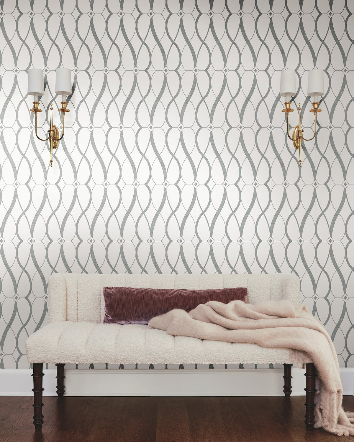Sure! Here’s your revised sentence incorporating the product data: A tufted cream bench with a mauve pillow and folded throw blanket sits in front of the Graceful Geo Wallpaper by York Wallcoverings. Two brass wall sconces with white lampshades are mounted above the bench, while a dark wooden floor grounds the space.