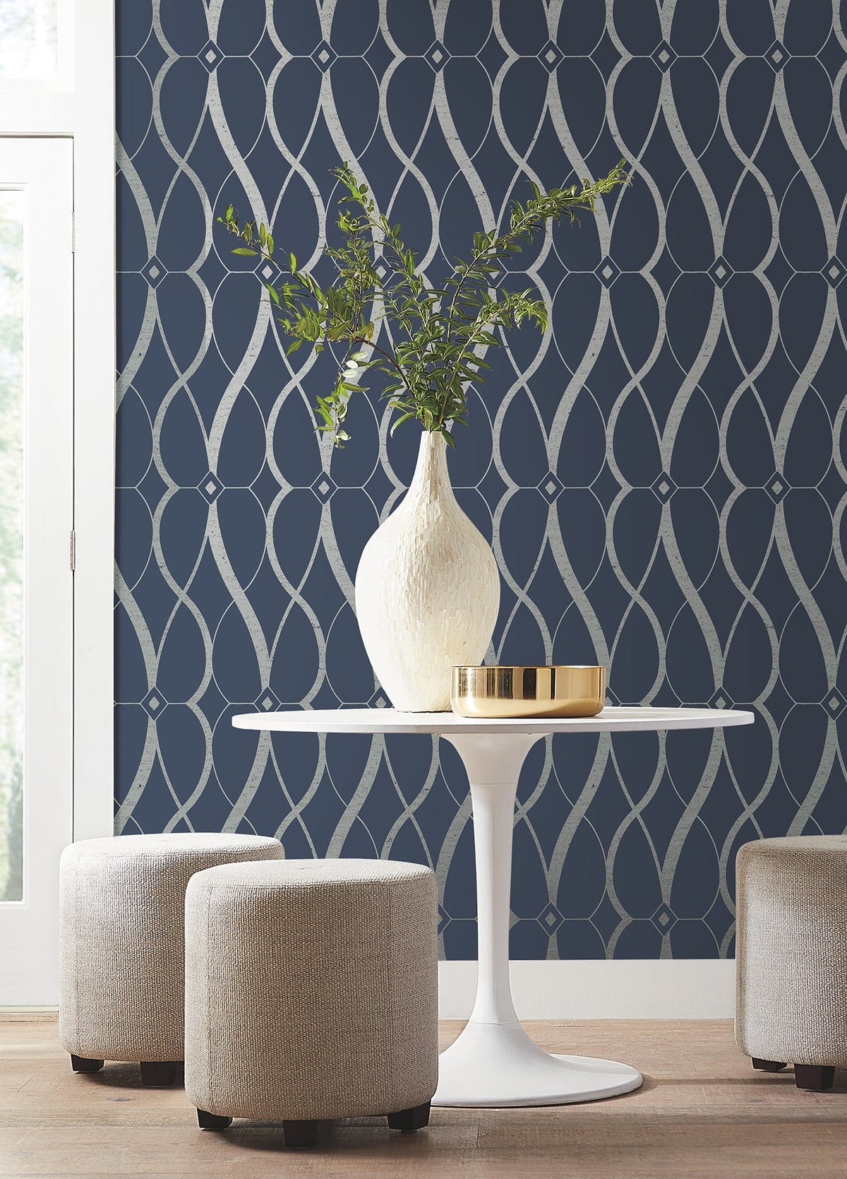 A round white table with a white vase of green branches and a gold tray, surrounded by three beige stools, sits in front of a navy accent wall adorned with York Wallcoverings’ Graceful Geo Wallpaper.
