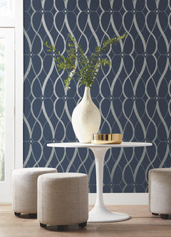 A round white table with a white vase of green branches and a gold tray, surrounded by three beige stools, sits in front of a navy accent wall adorned with York Wallcoverings’ Graceful Geo Wallpaper.
