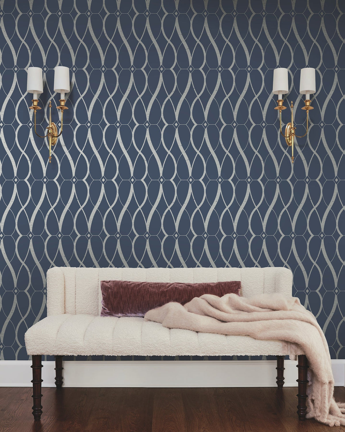 A cream-colored couch with a mauve pillow and a light pink throw sits against a navy wall adorned with York Wallcoverings’ Graceful Geo Wallpaper in a silver geometric pattern. Two gold wall sconces with white shades are mounted above the couch.